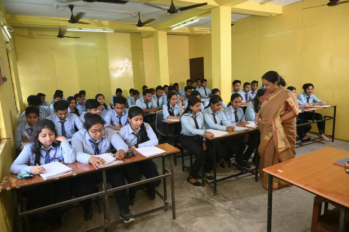 Students in uniform writing at their desks while a teacher walks between the rows